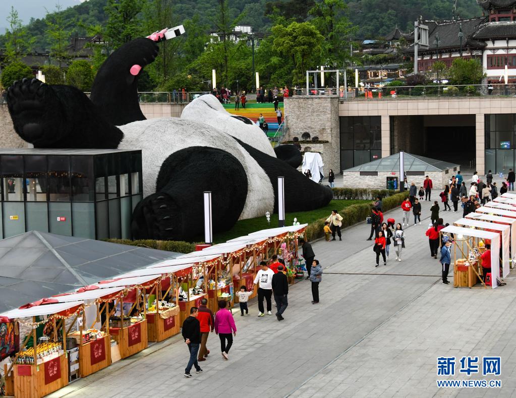 　　4月7日，在四川省都江堰市仰天窩廣場(chǎng)，游客在“自拍熊貓”雕塑旁邊的文創(chuàng)集市消費(fèi)。　　近日，由荷蘭藝術(shù)家弗洛倫泰因·霍夫曼設(shè)計(jì)的大熊貓自拍藝術(shù)品雕塑在四川省都江堰市仰天窩廣場(chǎng)與公眾見(jiàn)面。這只仰臥翹腳自拍的“大熊貓”長(zhǎng)26.5米、寬11米、高12米，重達(dá)130噸，由300萬(wàn)根漆面不銹鋼絲固定而成?！　?jù)了解，這只熊貓雕塑以2005年大熊貓闖入都江堰鬧市區(qū)的故事為創(chuàng)作靈感。熊貓手持一部巨型智能手機(jī)，吸引市民和游客前來(lái)觀賞，合影“打卡”?！　⌒氯A社記者 王曦 攝